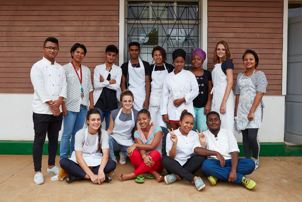 Gruppenfoto der Volontärinnen mit madagassischen Studenten vor einem Gebäude. Alle tragen Kochschürzen