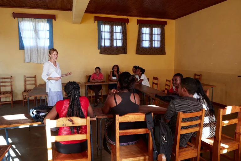 Eine kleine Gruppe junger Madagassen wird von einer Lehrerin unterrichtet. Das Klassenzimmer ist gelb gestrichen und die Sonne scheint durch die Fenster