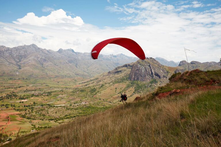 Ein Paraglider mit rotem Schirm kurz vor dem Abheben Richtung Tal. Im Hintergrund sind Berge zu sehen